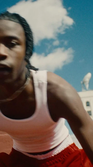 Close-up of Black man with braids in a white tank top against a bright blue sky and building with satellite dish.