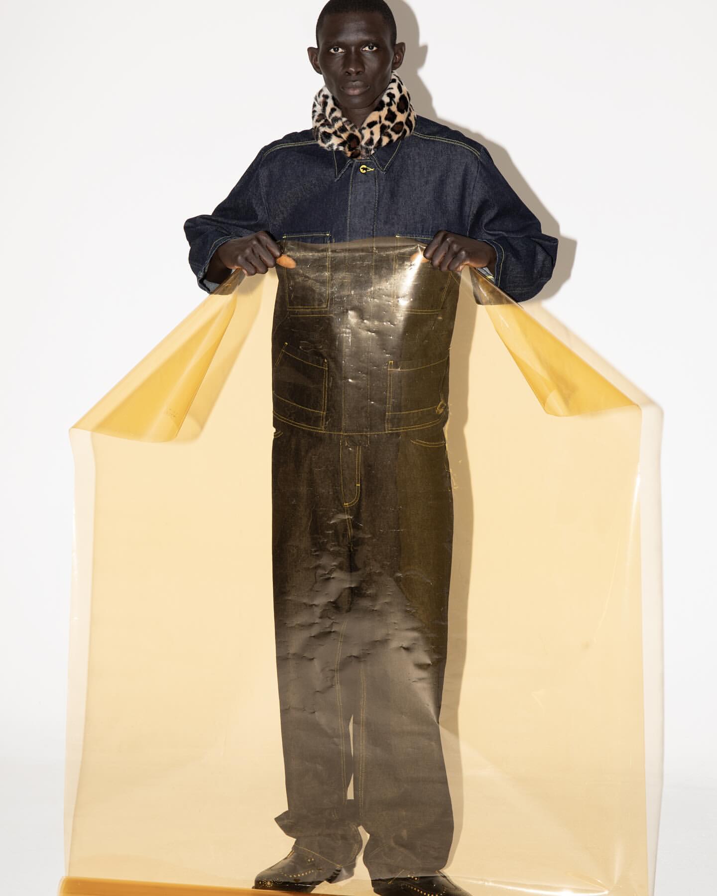 A person in denim with a scarf poses behind transparent material, creating a modern fashion portrait in a light studio setting.