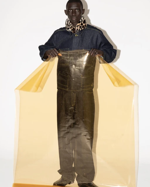 A person in denim with a scarf poses behind transparent material, creating a modern fashion portrait in a light studio setting.