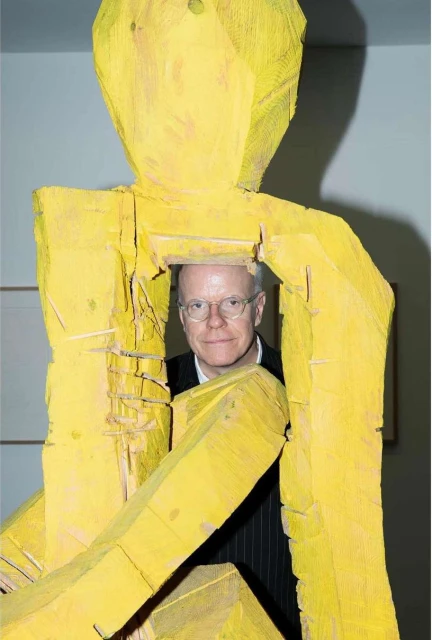 Adult male with glasses stands behind a yellow wooden statue, posing in an indoor setting with neutral walls and a dark suit.
