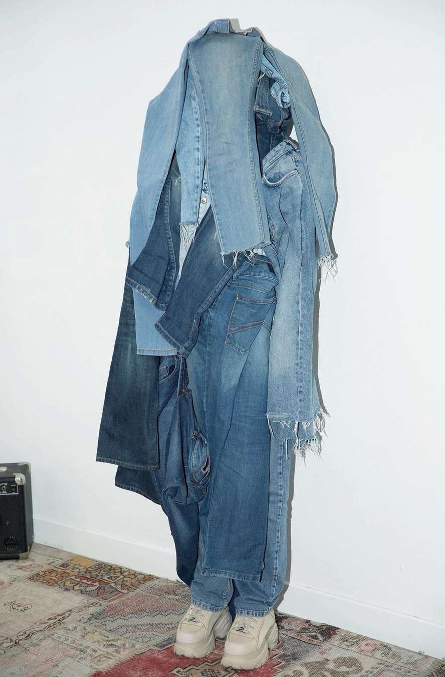 Full shot of a person covered in an array of blue denim jeans and white platform shoes, standing indoors on a patterned rug against a white wall.
