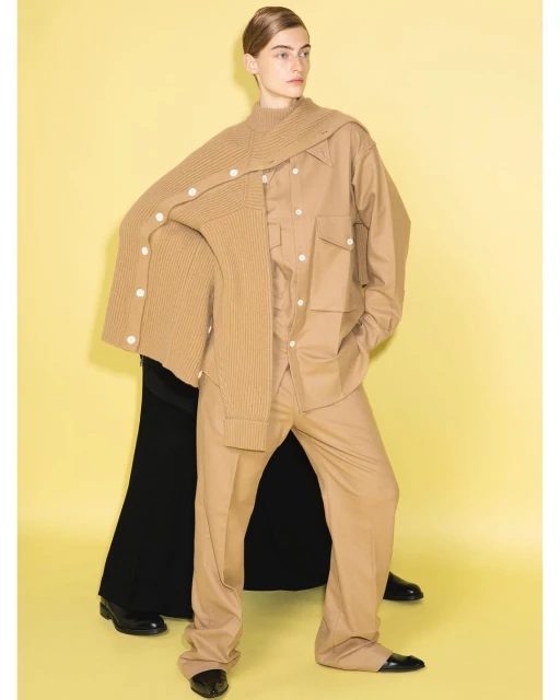 Two fashion models pose in a photo studio with a yellow backdrop, wearing beige and black outfits and shiny black shoes.