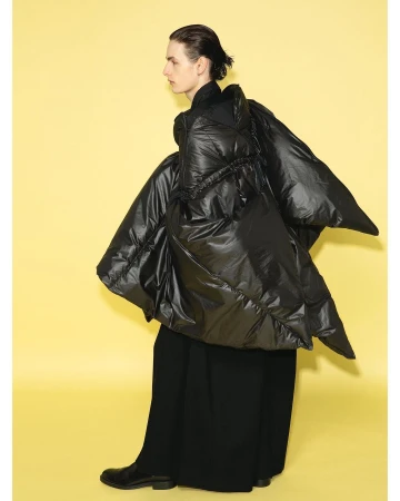 Side-view portrait of a person wearing a black puffer coat and long black skirt, posing against a light yellow backdrop in a studio.