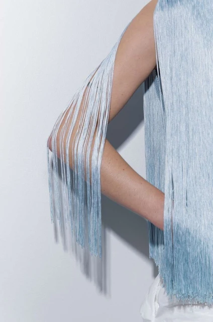 Close-up of a woman's arm and shoulder wearing a light blue fringe top, set against a neutral white background in a studio setting.