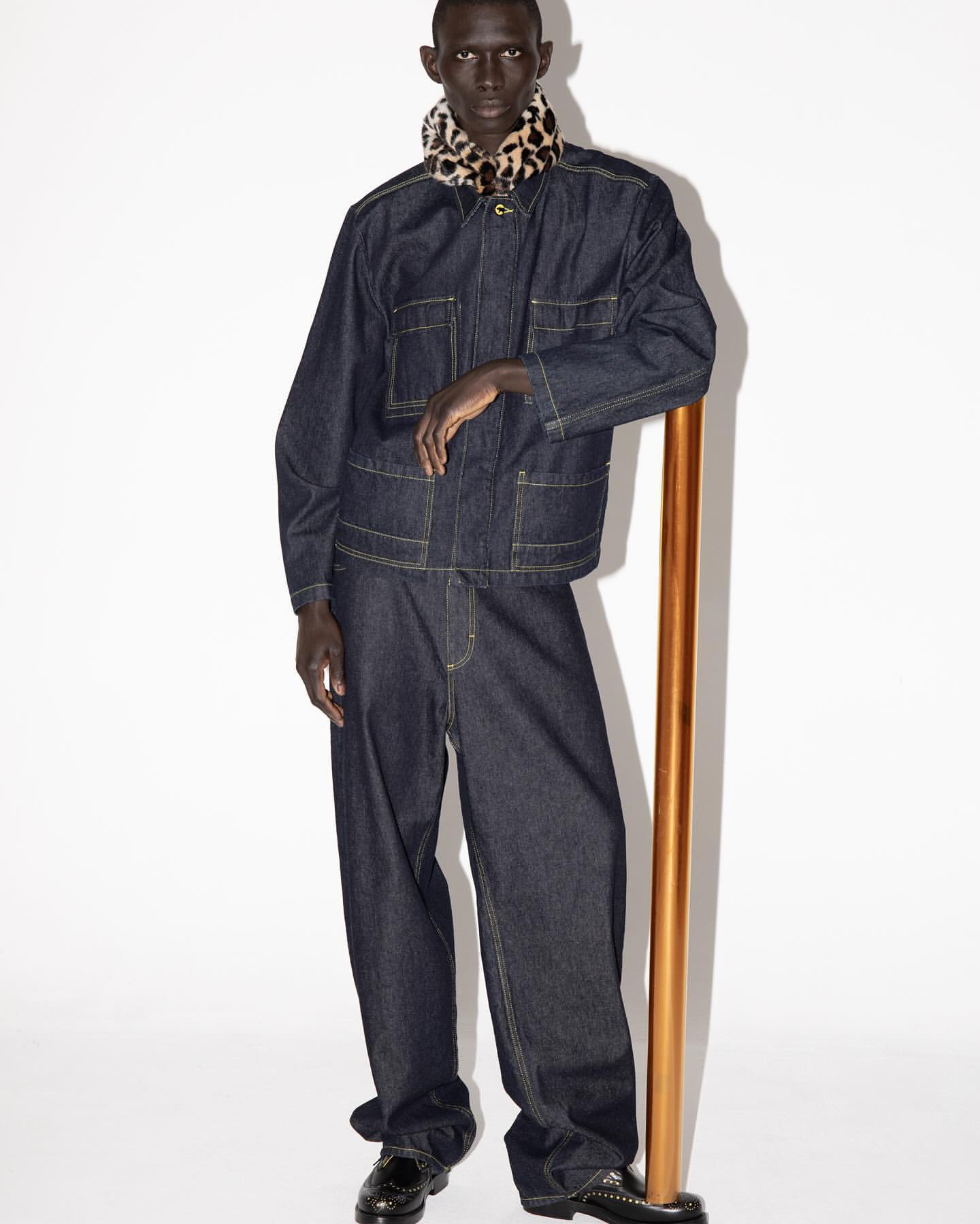 Studio shot of a Black male model in a dark denim jacket and pants with a cheetah print scarf and black dress shoes, leaning on a gold bar.