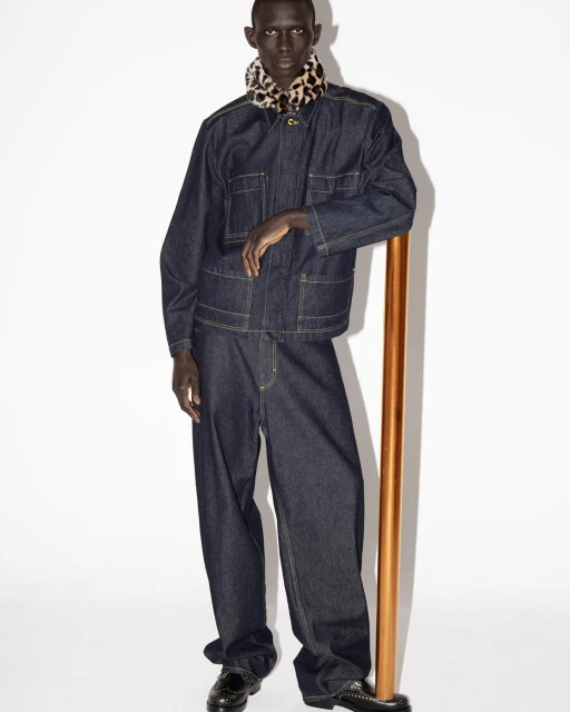 Studio shot of a Black male model in a dark denim jacket and pants with a cheetah print scarf and black dress shoes, leaning on a gold bar.