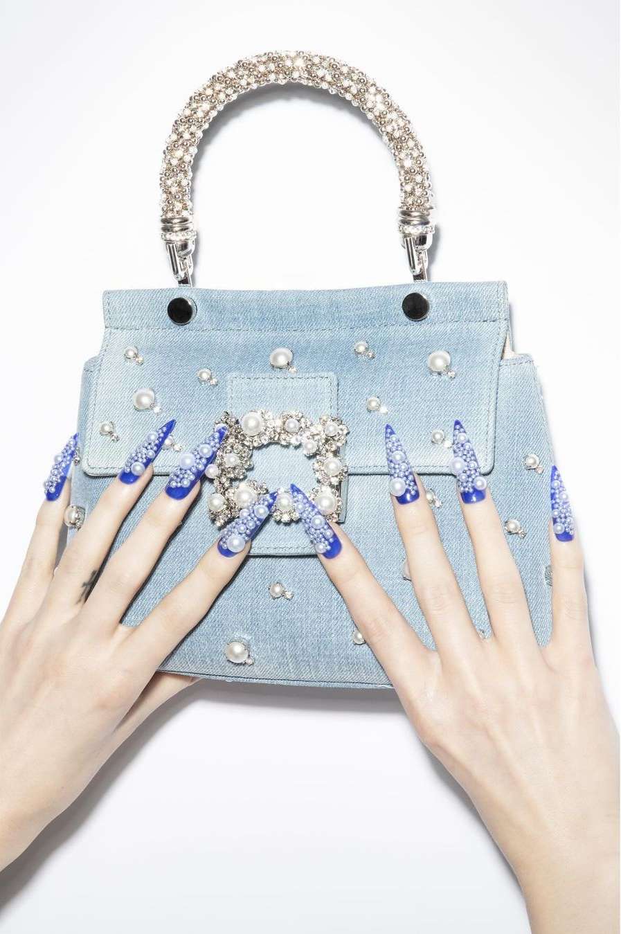 Fashion image of a light blue denim purse with pearl embellishments, styled with an adult female's hands featuring blue and white jeweled nails.
