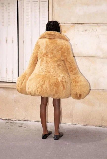 Person from the back wearing oversized furry tan coat outside against building with a door and concrete ground and light shining on ground.