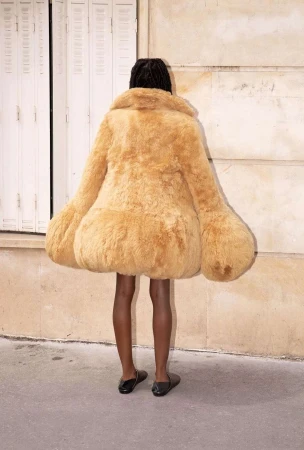 Person from the back wearing oversized furry tan coat outside against building with a door and concrete ground and light shining on ground.