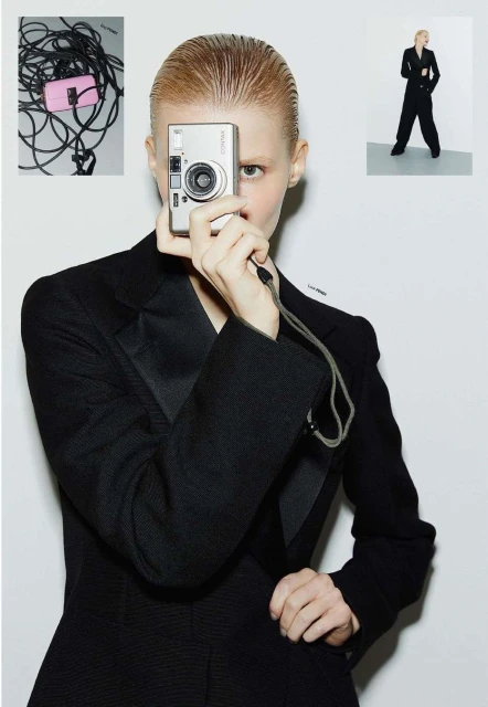 Adult woman in black suit holding silver camera in front of her face in a photo studio with other photography equipment visible.