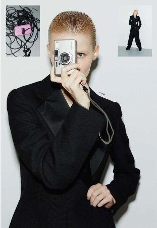 Adult woman in black suit holding silver camera in front of her face in a photo studio with other photography equipment visible.