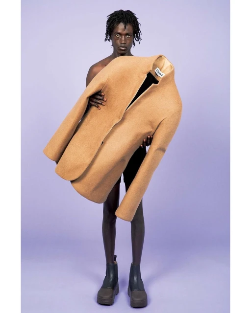An adult male holds an oversized tan blazer in a fashion forward pose against a lavender backdrop in a studio setting, emphasizing modern style.