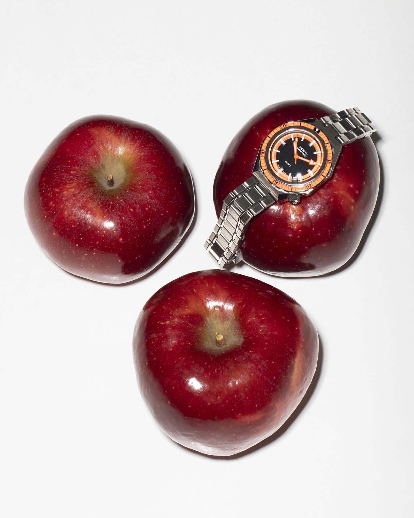 Three red apples arranged with a stainless steel wristwatch resting on one, shot in a studio with soft, diffused lighting.
