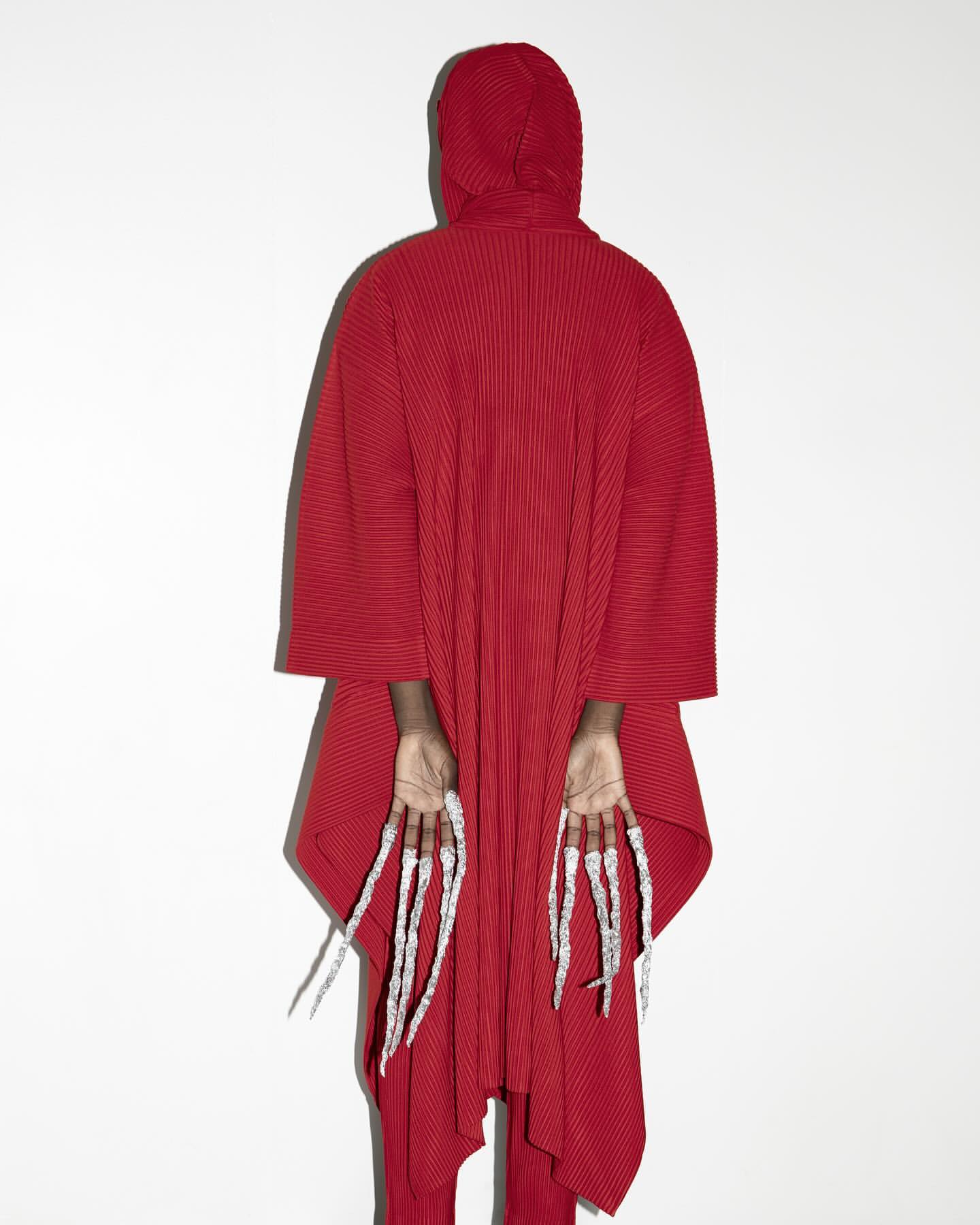Person in red hooded robe facing away, showing extra-long fingernails, against a white background. Fashion and beauty concept.