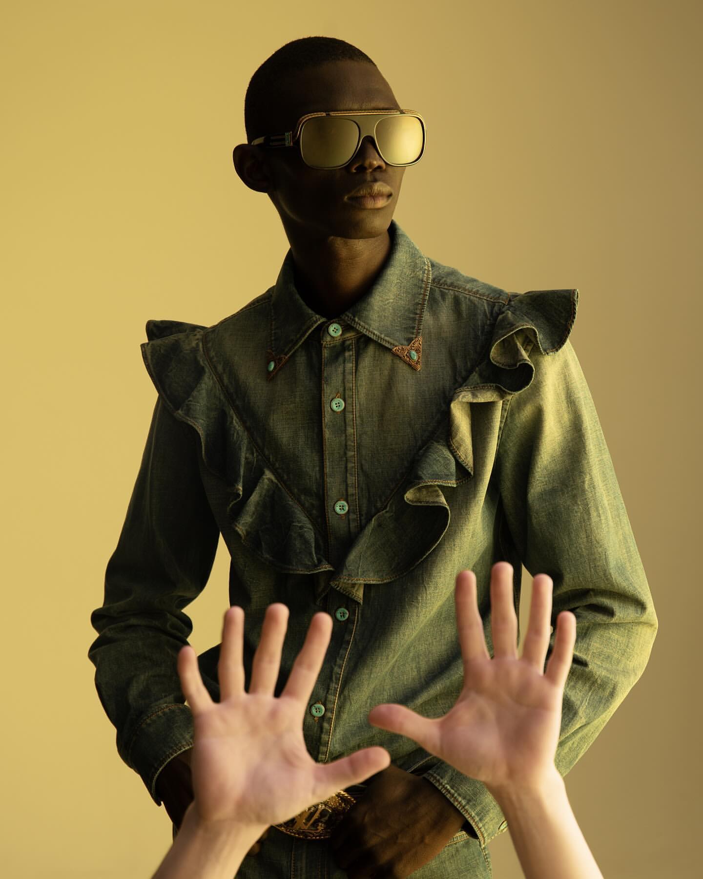 Black man in aviator sunglasses and ruffled denim shirt poses indoors, with hands partially obscuring the view. Fashion editorial shot.