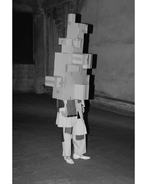 Person in cardboard box costume in a stark room. Black and white image of a unique avant-garde style with architectural backdrop.