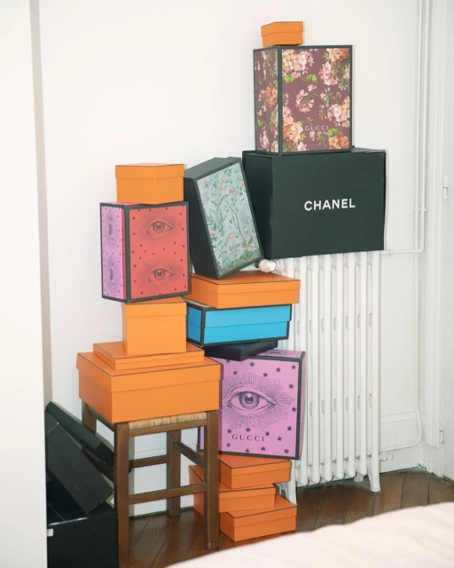 Stack of designer brand boxes, including Chanel and Gucci, on a wooden stool against a white wall indoors, fashion and luxury concept.