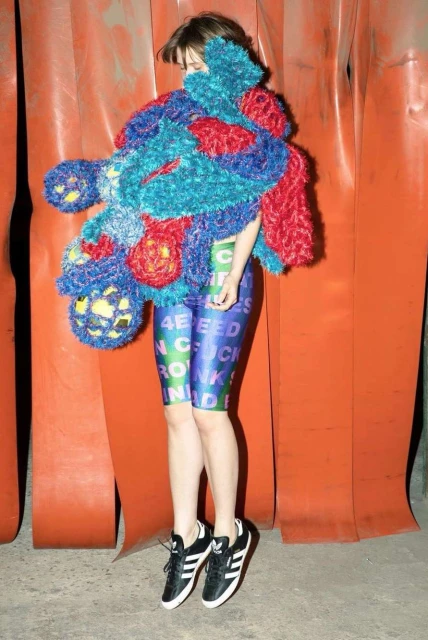 A young woman in colorful patterned leggings and sneakers stands against an orange background, partially covered by a fuzzy, vibrant shawl.