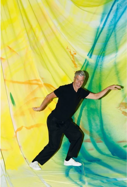 An adult male in a black shirt and pants poses dynamically in front of an abstract, brightly colored yellow, green, and blue backdrop.