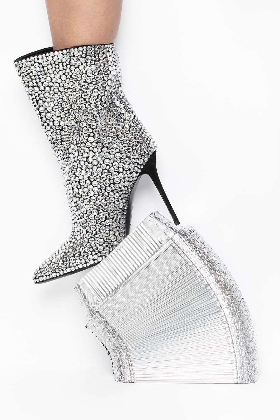 Rhinestone-covered ankle boot with a black heel, angled next to stacked silver fan blade covers on a white backdrop. Fashion footwear.