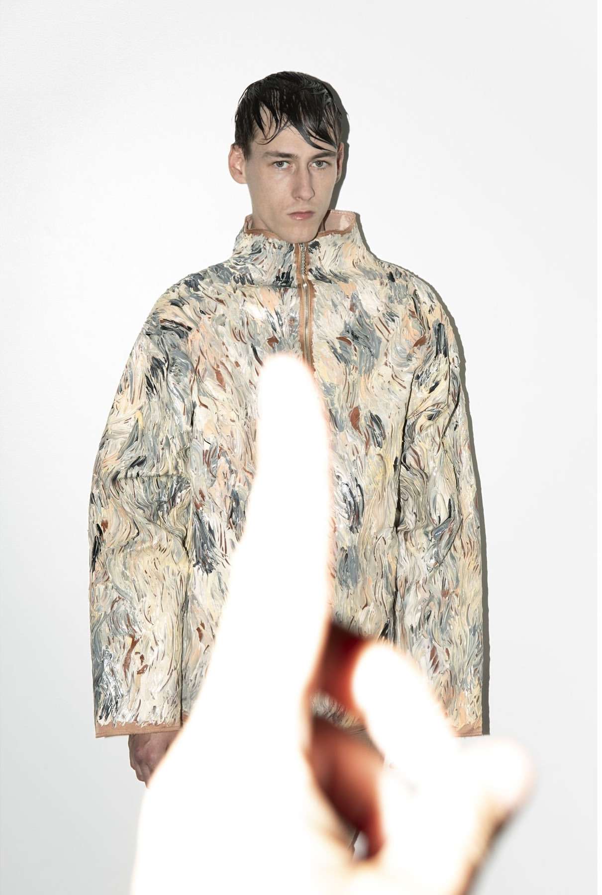 Male model in abstract-patterned jacket in a studio with an out-of-focus hand pointing up from the foreground, against a white background.