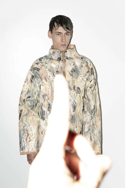 Male model in abstract-patterned jacket in a studio with an out-of-focus hand pointing up from the foreground, against a white background.