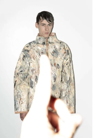 Male model in abstract-patterned jacket in a studio with an out-of-focus hand pointing up from the foreground, against a white background.