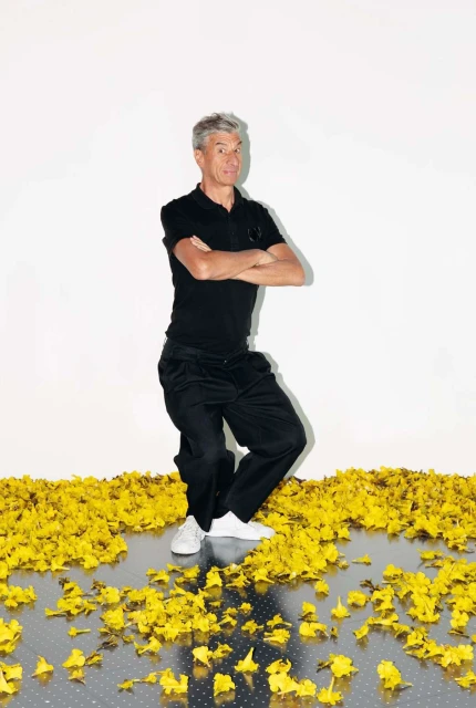 Full shot of a mature adult man with grey hair standing on yellow flower petals in a black polo shirt and pants, against a white background.