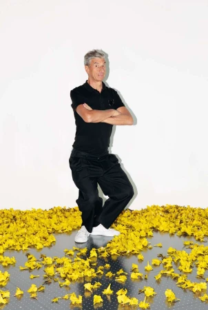 Full shot of a mature adult man with grey hair standing on yellow flower petals in a black polo shirt and pants, against a white background.