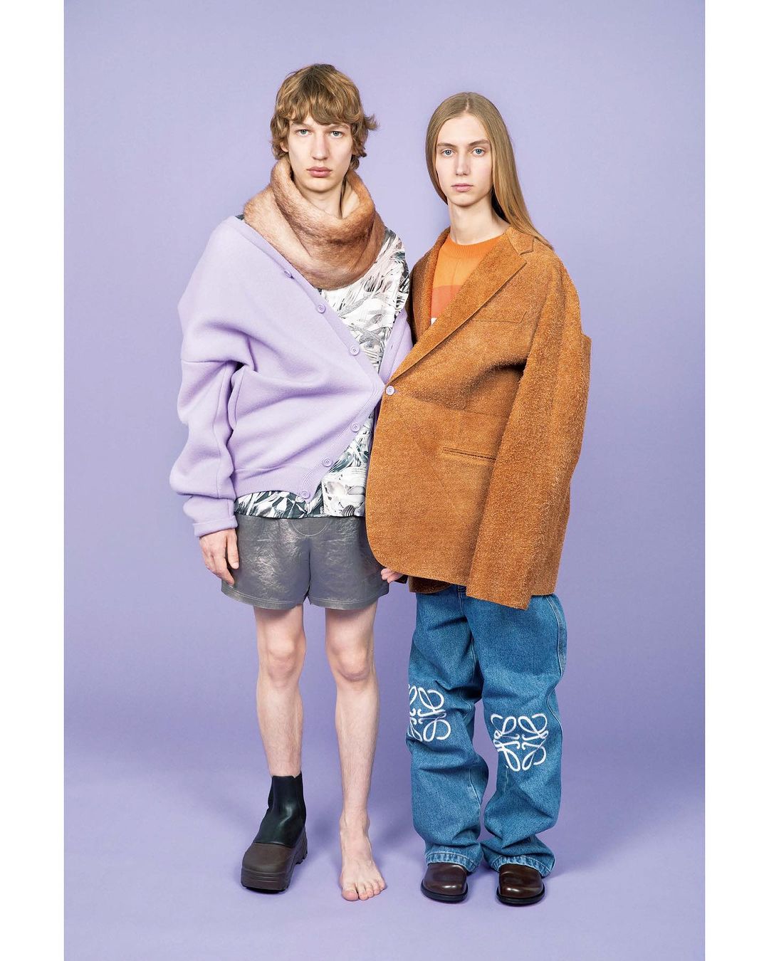 Two models in avant-garde fashion stand against a solid lavender backdrop, featuring a mix of textures and oversized garments.