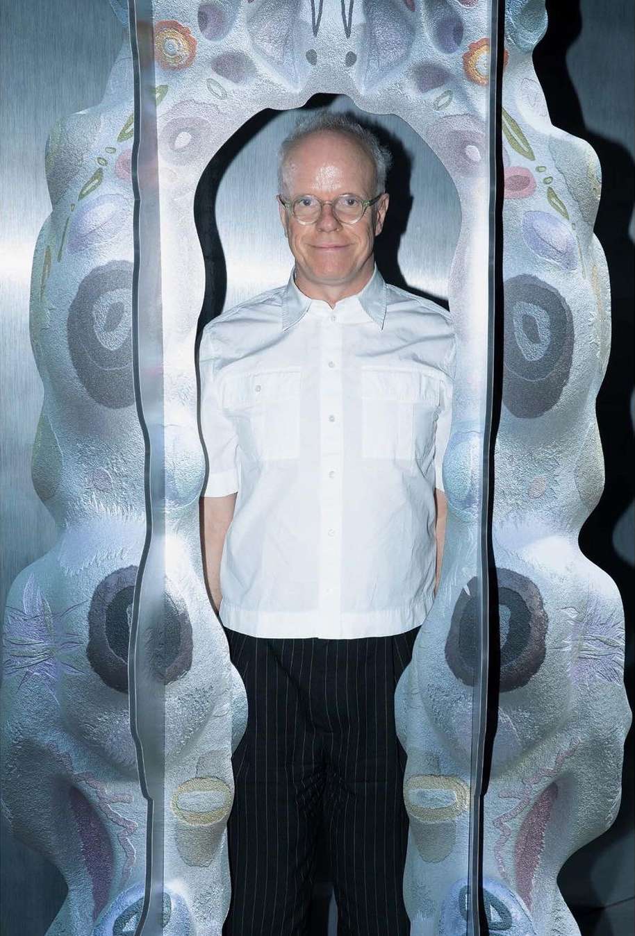 Adult male with glasses and white shirt standing inside of an open art sculpture in an indoor setting with studio lighting. 