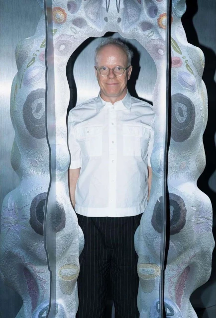 Adult male with glasses and white shirt standing inside of an open art sculpture in an indoor setting with studio lighting. 
