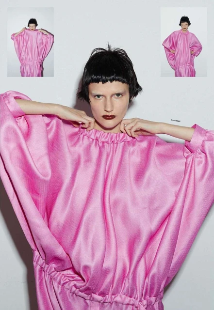 Fashion shot of an adult female model wearing a pink dress with hands near her neck, set against a bright white studio backdrop.