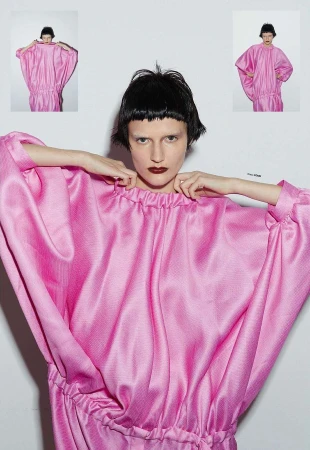Fashion shot of an adult female model wearing a pink dress with hands near her neck, set against a bright white studio backdrop.