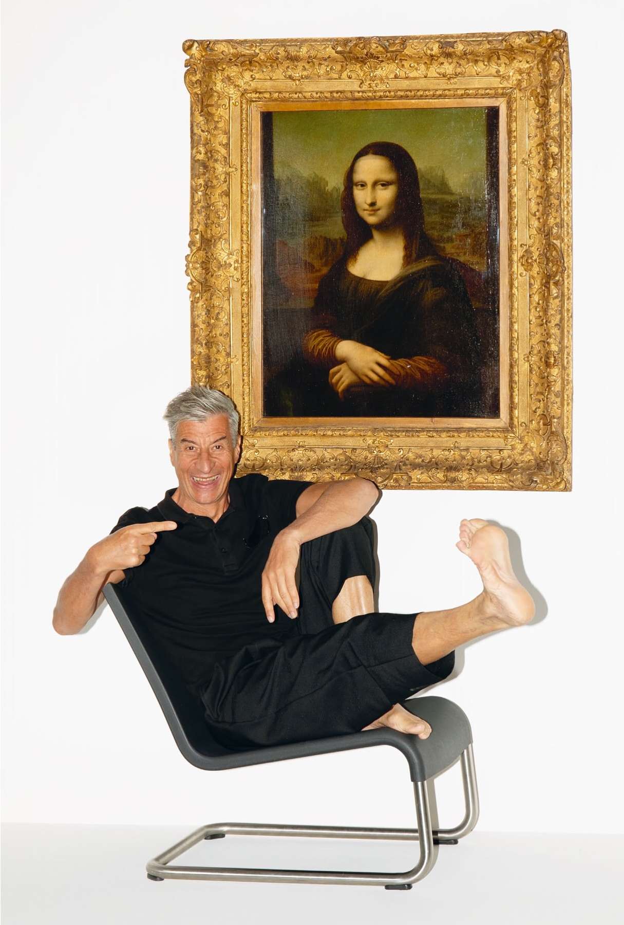 Mature man poses in a chair pointing towards a gold-framed portrait of the Mona Lisa, set against a stark white backdrop.