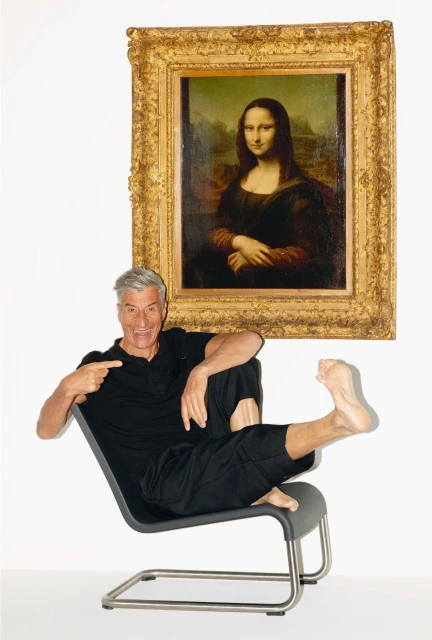 Mature man poses in a chair pointing towards a gold-framed portrait of the Mona Lisa, set against a stark white backdrop.