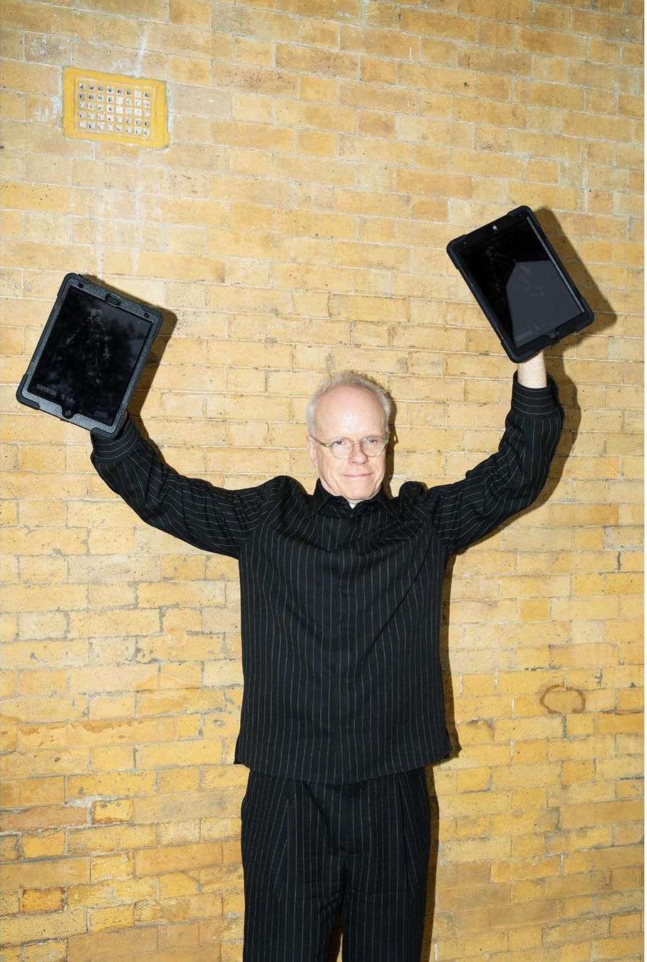 Older man in pinstripe suit, raising two tablets against a brick wall background, creating a balanced and tech-forward image.