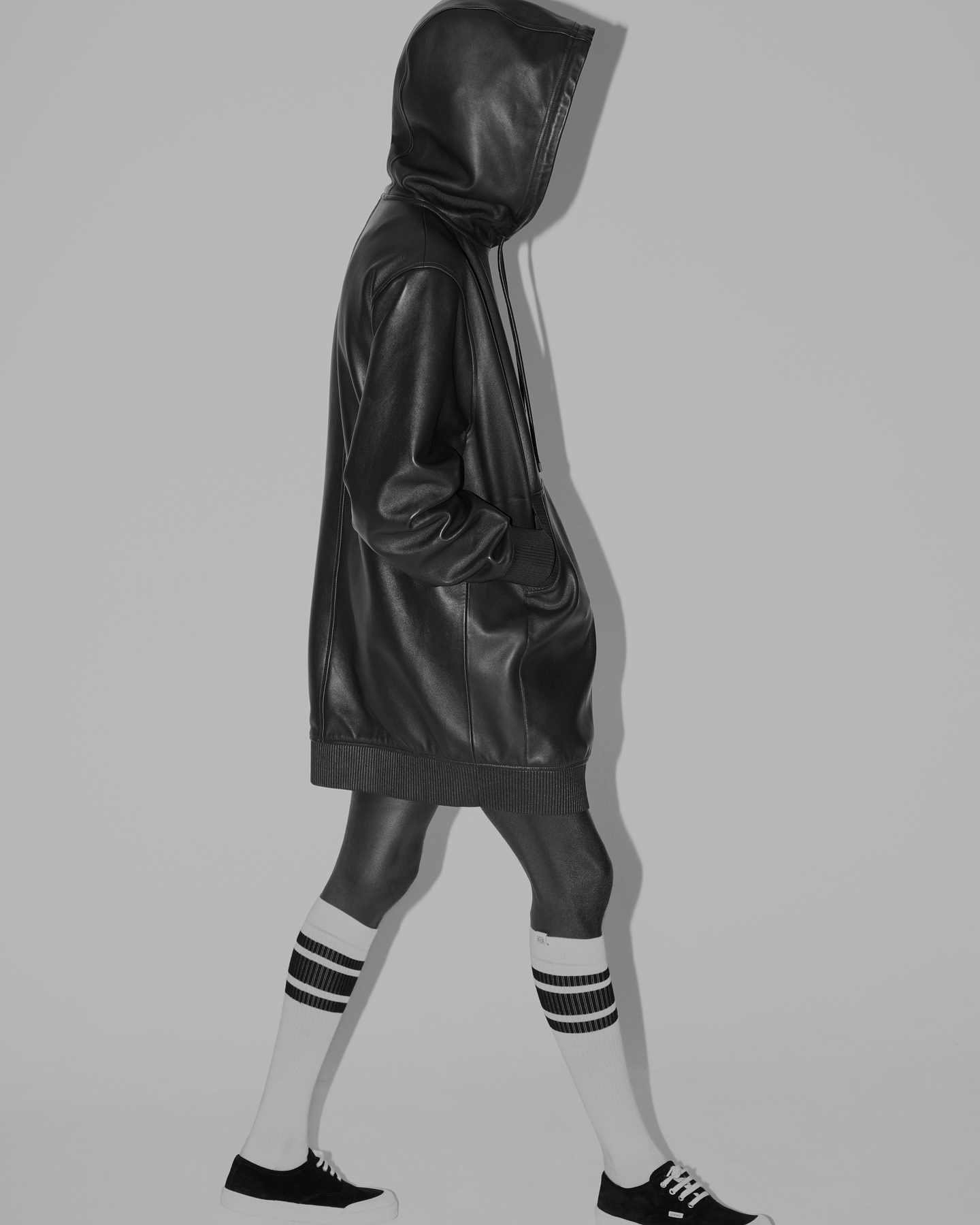 Person in black leather hoodie and black and white striped socks walking in a studio setting against a neutral grey backdrop.