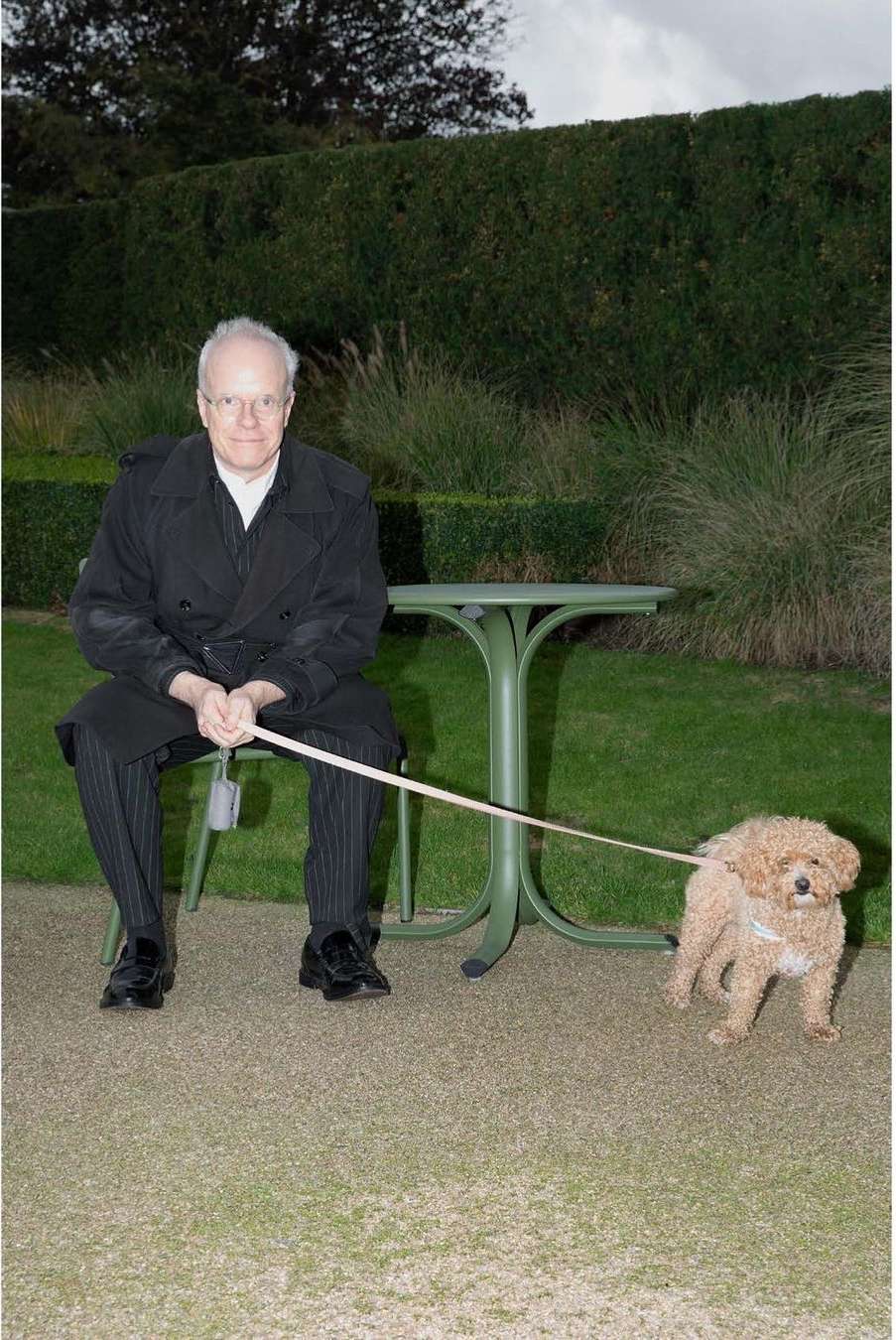 Adult man sits outdoors holding a leash attached to a poodle; table and hedge in background, in a full shot.