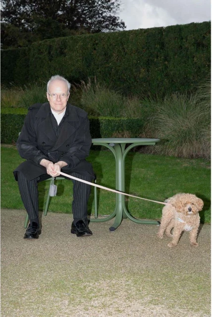 Adult man sits outdoors holding a leash attached to a poodle; table and hedge in background, in a full shot.