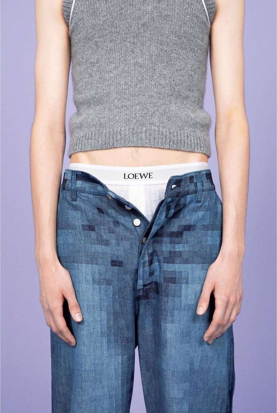Fashion shot featuring a slender adult in a grey top and blue patterned pants, revealing white underwear against a light purple backdrop.