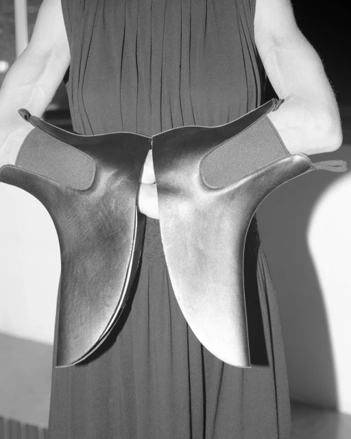 Black and white studio shot of person holding pair of wide, dark-colored protective leg coverings for horse riding in front of them.