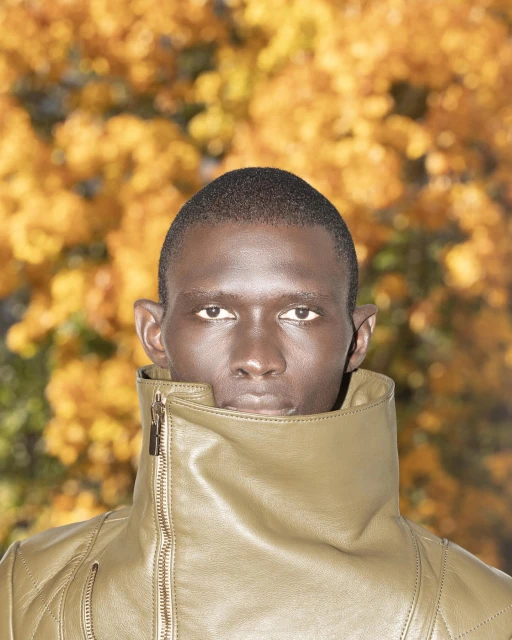 Person in olive green, high-collared, quilted jacket poses outdoors with blurred golden autumn foliage in the background.