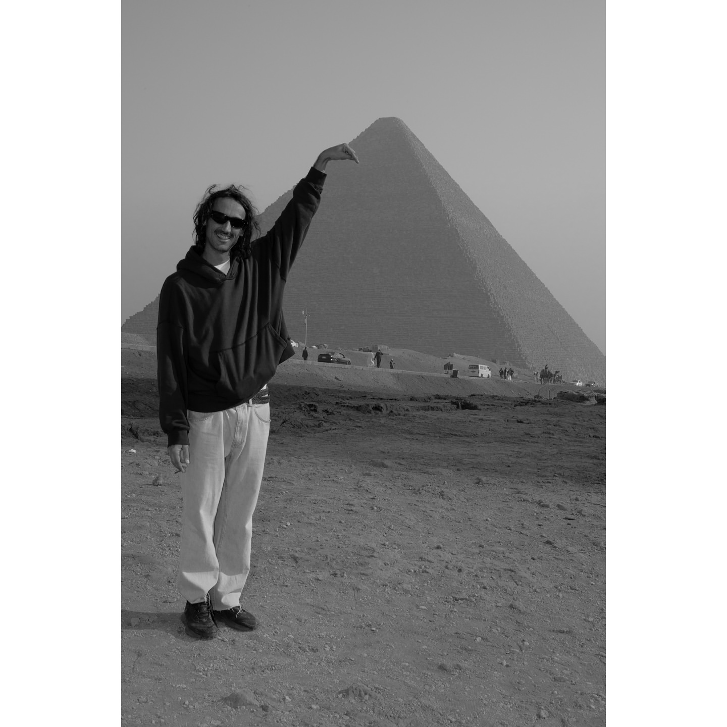 Monochrome full shot of a man in casual wear posing with arm raised near the Great Pyramid of Giza, Egypt, under a clear sky.