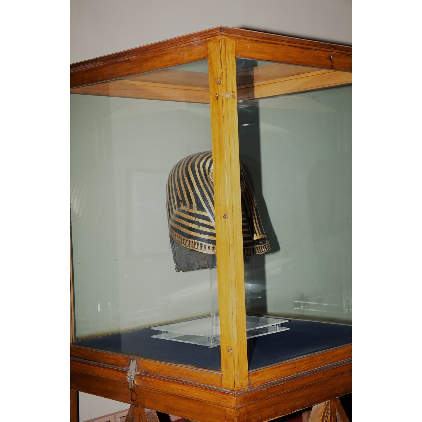 Ancient Egyptian helmet displayed inside a wooden and glass museum display case, showcasing intricate gold stripes and craftsmanship.