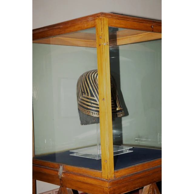 Ancient Egyptian helmet displayed inside a wooden and glass museum display case, showcasing intricate gold stripes and craftsmanship.