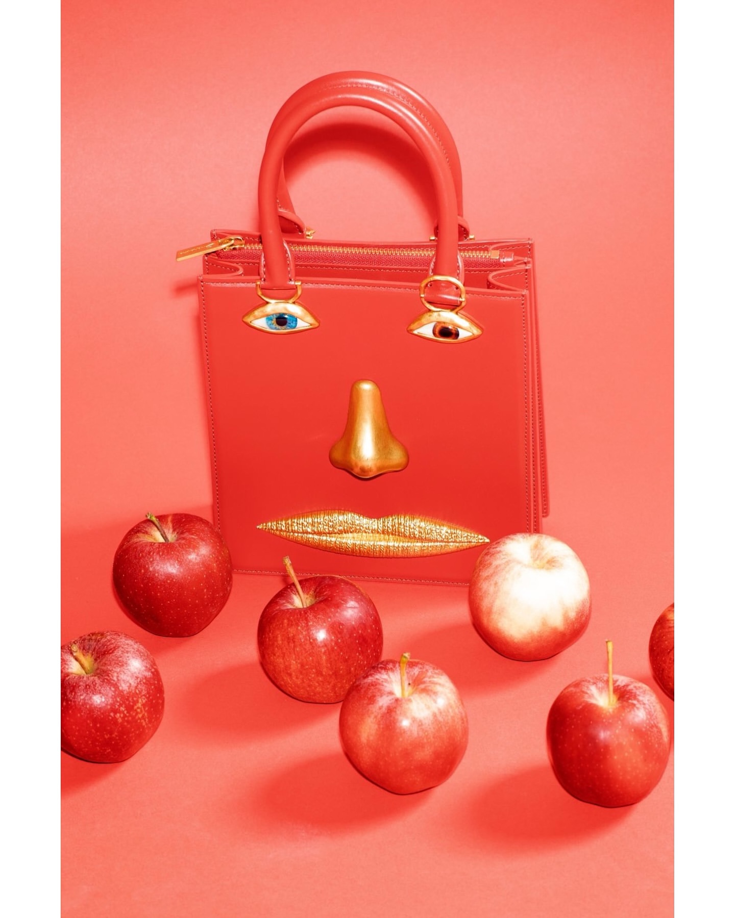 Coral handbag with whimsical eye, nose, and lip appliques surrounded by red apples against a matching coral background.