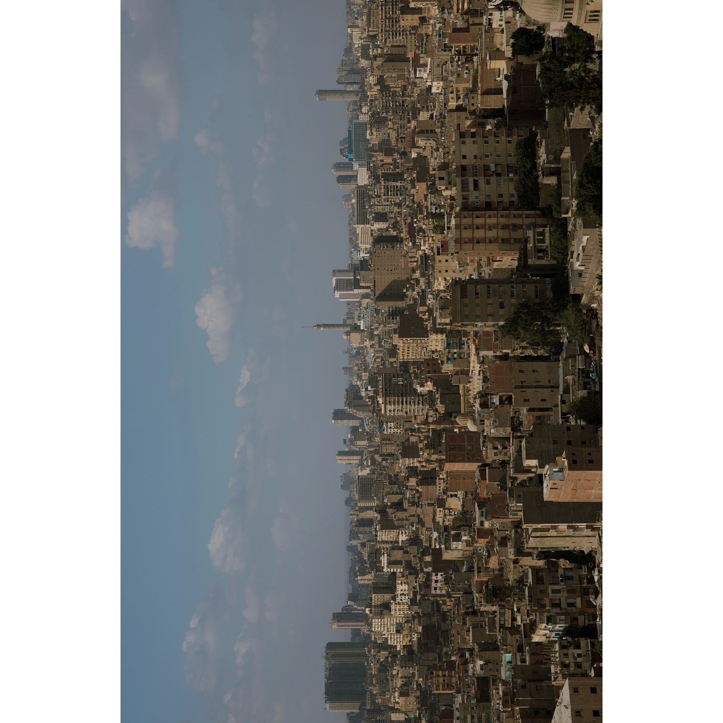 Cityscape aerial view featuring dense buildings, a tall tower, and a hazy blue sky with scattered clouds, showcasing urban architecture.