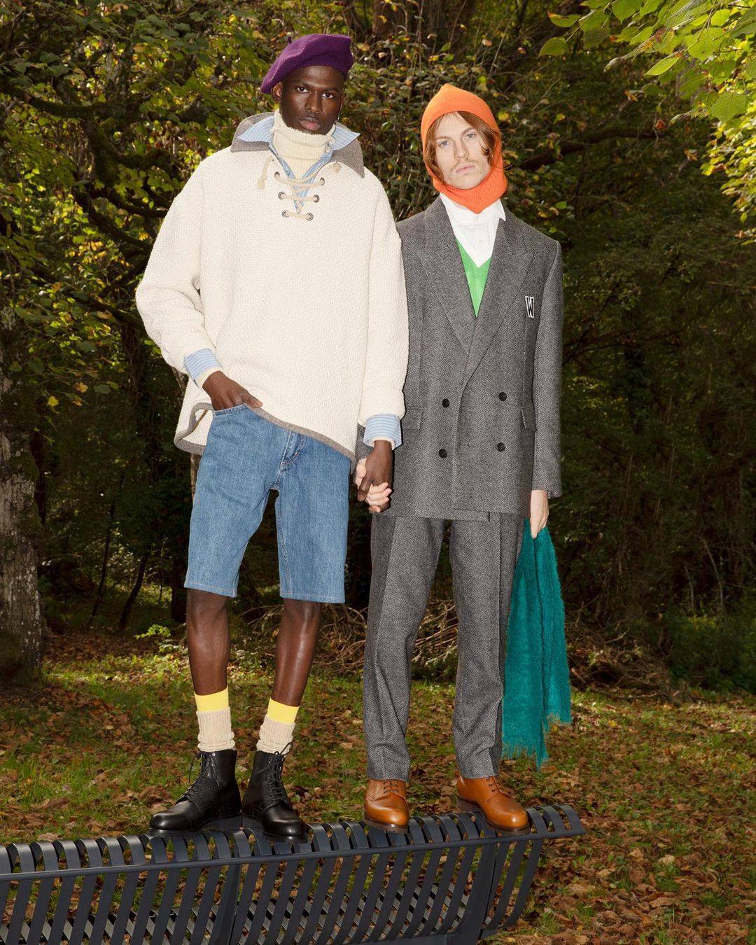 Two male models holding hands standing on a black metal park bench in a wooded setting, captured by Jonathan Llense in Paris.