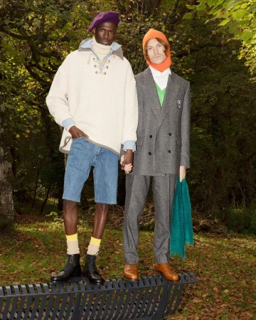Two male models holding hands standing on a black metal park bench in a wooded setting, captured by Jonathan Llense in Paris.